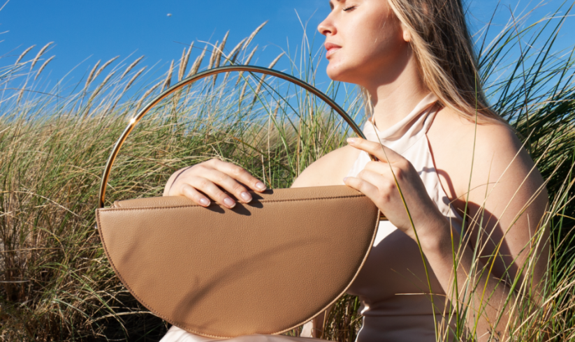 A model showcasing a Venus & Aphrodite quiet luxury bag in light tones on a summer day in the grass, embodying elegance and versatility.